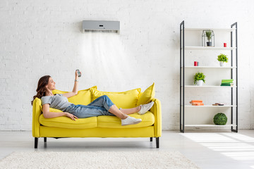 attractive young girl lying on yellow sofa under air conditioner at home - obrazy, fototapety, plakaty