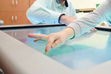 The hand of the child stretches to press the icon on the touchscreen of the interactive table