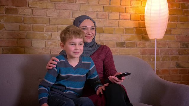 Portrait of small boy and his muslim mother in hijab watching TV together sitting on sofa at home.