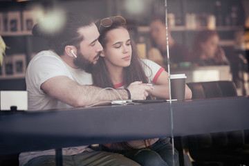 View through the glass nice young couple in love chatting and looking at each other sit at a table in a cafe with a cup of coffee and a smartphone. Concept free high-speed wi-fi