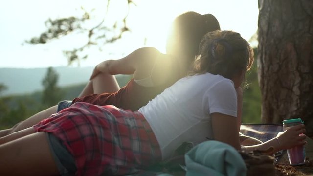Slow-motion two beautiful girls contemplating breathtaking peaceful view lying picnic blanket drink coffee metal mug look sunset explore countryside hiking mountain range, travelling together.