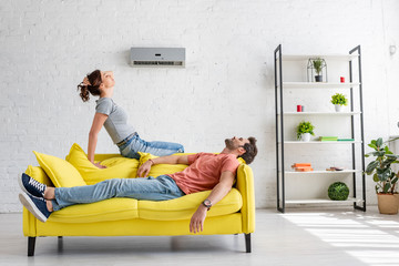 young man and woman resting on yellow sofa under air conditioner at home - obrazy, fototapety, plakaty