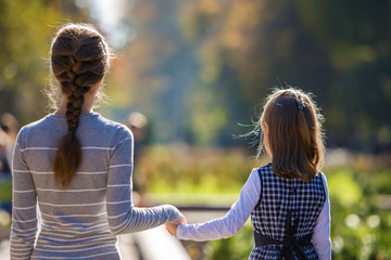 Back view of child girl and mother in dresses together holding hands on warm day outdoors on sunny background.
