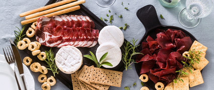 banner of cold cuts and cheese are served on a tray on a table with white wine, crackers, grissini and taralli with aromatic herbs on a blue linen festive tablecloth.