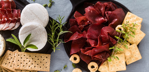 banner of Italian bresaola served sliced on a tray on a table with white wine, crackers, grissini and taralli with aromatic herbs on a blue linen festive tablecloth.