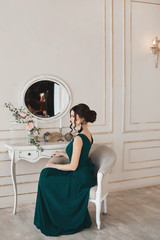 Fashionable young model woman with stylish hairstyle in trendy evening dress sits in the armchair near the dressing table
