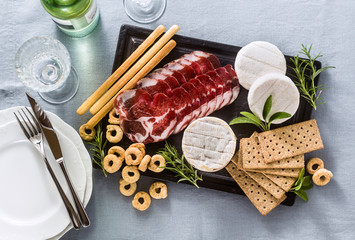 cold cuts and cheese are served on a tray on a table with white wine, crackers, grissini and taralli with aromatic herbs on a blue linen festive tablecloth.