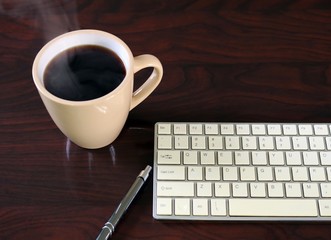 Steaming hot cup of coffee beside keyboard on dark wood grain desk