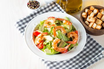 Fresh shrimp salad made of tomato, ruccola, avocado, prawns, chicken breast, arugula, crackers and spices. Caesar salad in a white, transparent bowl on wooden background