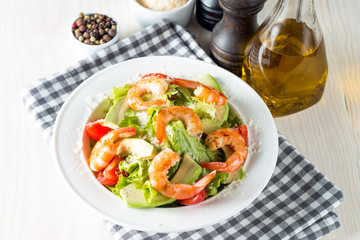 Fresh shrimp salad made of tomato, ruccola, avocado, prawns, chicken breast, arugula, crackers and spices. Caesar salad in a white, transparent bowl on wooden background