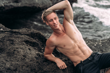 Fototapeta na wymiar Portrait of young man with naked torso standing on rock beach with black sand
