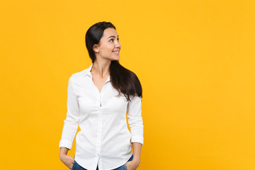 Portrait of smiling young woman in white shirt standing, holding hands in pockets, looking aside isolated on yellow orange wall background in studio. People lifestyle concept. Mock up copy space.