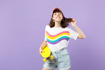Portrait of charming teen girl in vivid clothes with yellow skateboard holding hair isolated on violet pastel wall background in studio. People sincere emotions, lifestyle concept. Mock up copy space.