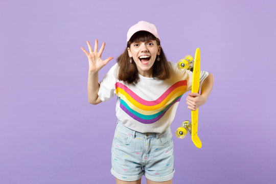 Portrait of cheerful teen girl in vivid clothes holding yellow skateboard, showing pelm isolated on violet pastel background in studio. People sincere emotions, lifestyle concept. Mock up copy space.