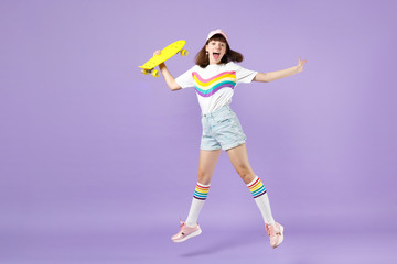 Excited teen girl in vivid clothes hold skateboard, having fun, jumping, keeping mouth open isolated on violet pastel wall background. People sincere emotions, lifestyle concept. Mock up copy space.