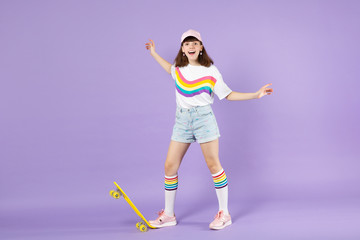 Cheerful teen girl in vivid clothes standing with yellow skateboard spreading hands isolated on violet pastel wall background in studio. People sincere emotions, lifestyle concept. Mock up copy space.