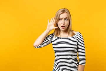Obraz na płótnie Canvas Curious young woman in striped clothes trying to hear you, holding hand near ear isolated on yellow orange wall background in studio. People sincere emotions, lifestyle concept. Mock up copy space.