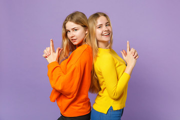 Two young blonde twins sisters girls in colorful clothes standing back to back, keeping fingers like gun isolated on pastel violet blue background. People family lifestyle concept. Mock up copy space.