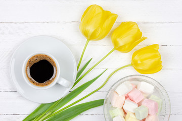 a Cup of black coffee on a saucer, a bouquet of yellow tulips and marshmallows on a white wooden background, top view flat lay