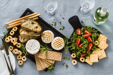 grilled Italian Tomino cheese served on a table with white wine, crackers, grissini and taralli with aromatic herbs and arugula and tomato salad on a blue linen festive tablecloth. summer menu
