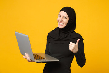 Young arabian muslim employee woman in hijab black clothes hold and work laptop pc computer isolated on yellow wall background, studio portrait. People religious lifestyle concept. Mock up copy space.