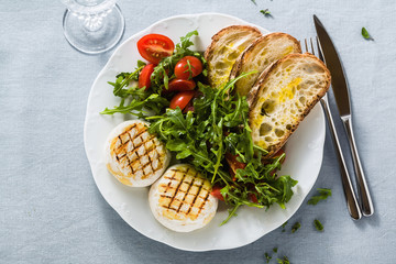 grilled Italian Tomino cheese served on a table with arugula salad and fresh homemade ciabatta bread and tomato on a blue linen festive tablecloth. summer menu