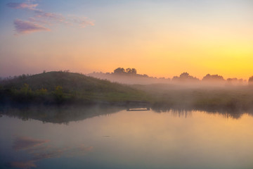 Fototapeta na wymiar Picturesque misty sunrise over a lake with a bench and a park