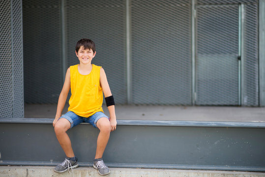 Front View Of Smiling Boy Sitting On A Metallic Fence While Looking To Camera On A Bright Day