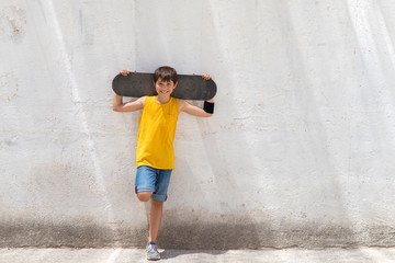 Young smiling boy leaning on yellow wall holding a skateboard while looking camera in a bright day