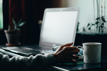 Young adult hand reaching for a white cup of coffee while working on a laptop with a green plant in background – Concept image for online shopping and working from home