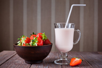 strawberry yogurt with a straw decorated with a strawberry in the cut and a bowl of strawberries on a brown wooden table, side view