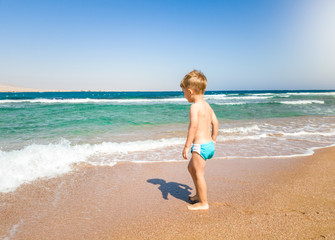 Little toddler boy walking on the beach and looking at calm and warm sea waves. Child relaxing and having good time during summer holiday vacation.