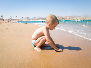 Adorable 3 years old toddler boy sitting on the sea beach in warm calm waves and playing with wet sand. Child relaxing and having good time during summer holiday vacation.