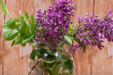 Fototapeta premium Fresh cut Purple Lilac Flowers in clear glass vase on wooden background. Syringa vulgaris.