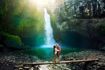 Young couple enjoy honeymoon vacation on tropical island Bali, hugs and kisses on background scenery waterfall Tegenungan and amazing nature landscape at sunrise - obrazy, fototapety, plakaty