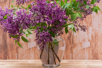Fresh cut Purple Lilac Flowers in clear glass vase on wooden background. Syringa vulgaris.
