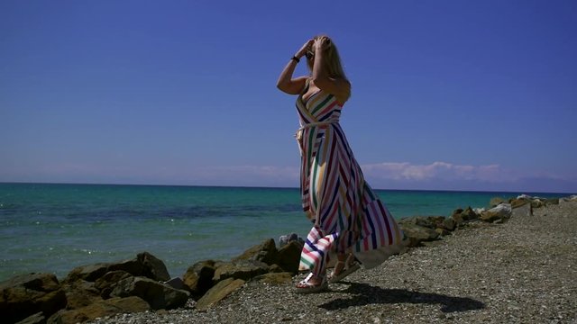 happy blonde woman wearing long striped dress is walking on edge of shore near sea