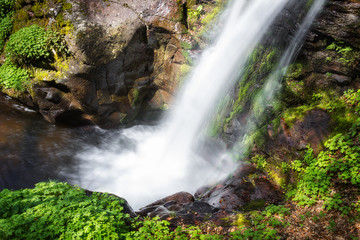 Naklejka na ściany i meble View from above of scenic forest waterfall Krmolj on old mountain, with sunlit rocks and plants vivid green during early spring, streaming down the vertical red cliff