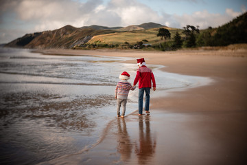 Two boys wearing Santa hats walking on a beach with mountains