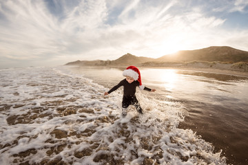 Fototapeta premium Toddler boy wearing red hat splashing in waves at beach at dusk