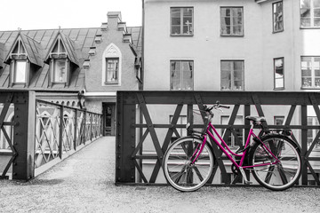 A picture of a lonely pink bike standing in the typical street in Stockholm by the bridge to a house. The bike looks to be modern in a retro style. The background is black and white. 