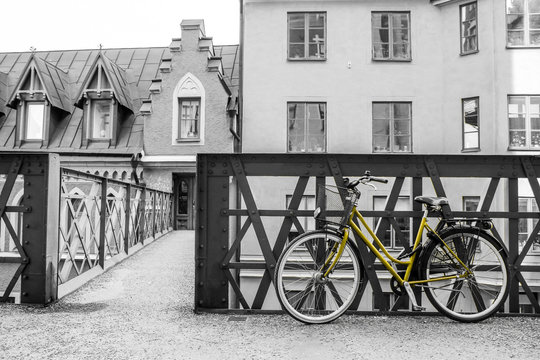 A picture of a lonely yellow bike standing in the typical street in Stockholm by the bridge to a house. The bike looks to be modern in a retro style. The background is black and white. 