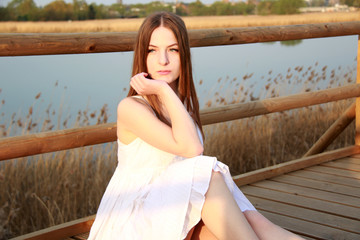 young girl in white dress sitting next to the lake on wooden bridge