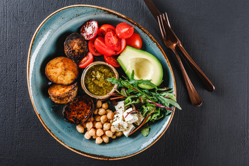 Fototapeta na wymiar Healthy Buddha bowl dish with avocado, tomato, cheese, chickpea, fresh arugula salad, baked potatoes and sauce pesto in black background. Dieting food, clean eating, top view.