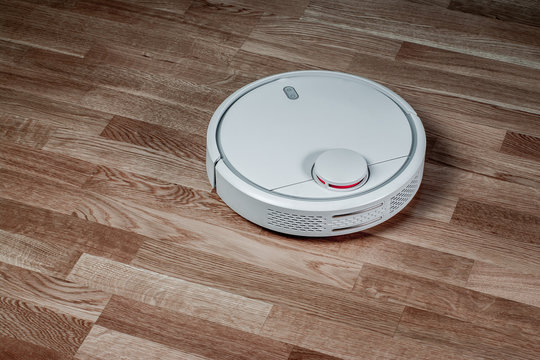 white robotic vacuum cleaner runs on laminate floor. Robot controlled by voice commands for direct cleaning. Modern smart appliance for cleaning house