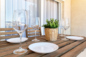 Close up white empty plates wine glasses, wooden table setting at home in outdoors terrace, plant on weave pot no people.