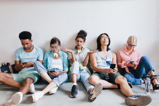 Bored girl with curly black hair angry beacause her college friends don't listen her. Tired international students sitting on the floor and using theis phones after lecture.