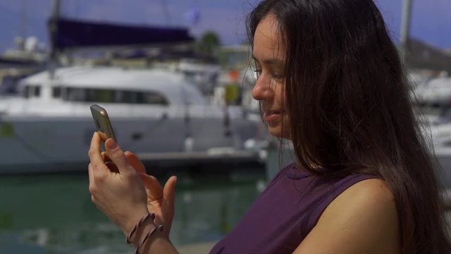 charming brunette woman is photographing herself by smartphone in pier with small yachts