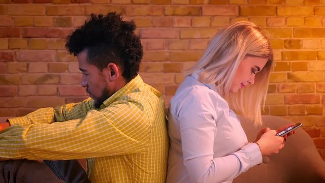 Close-up shot of african guy and caucasian girl sitting back to back on sofa watching into their smartphones at home.