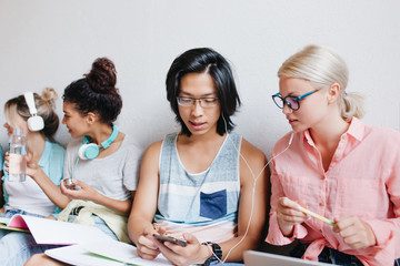 Indoor portrait of group of people with blonde girl and asian boy on foreground. Attractive fair-haired female student in elegant glasses enjoying music in earphones with university friend.